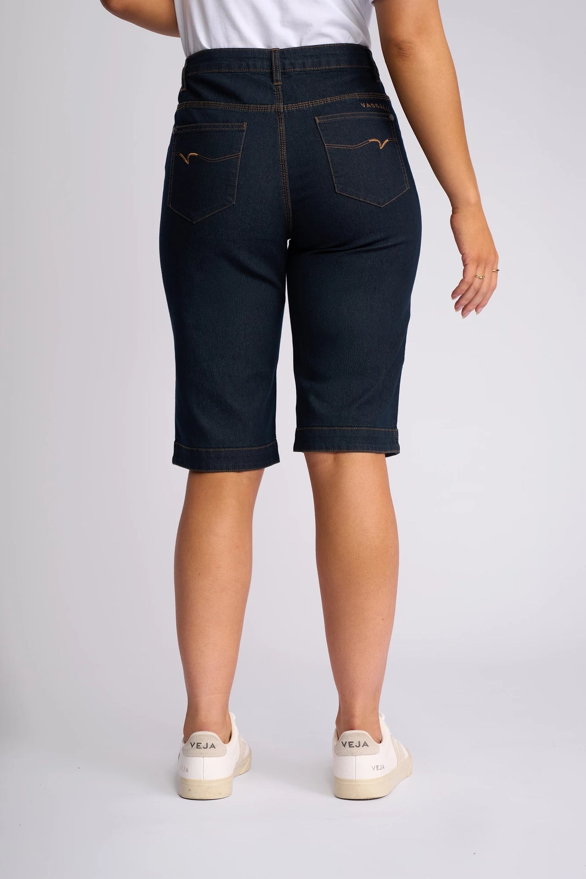 A person wearing the Vassalli Custom Short - Indigo Denim, paired with white Veja sneakers and a white top, stands with their back to the camera against a plain light background—effortlessly embracing summer dressing.