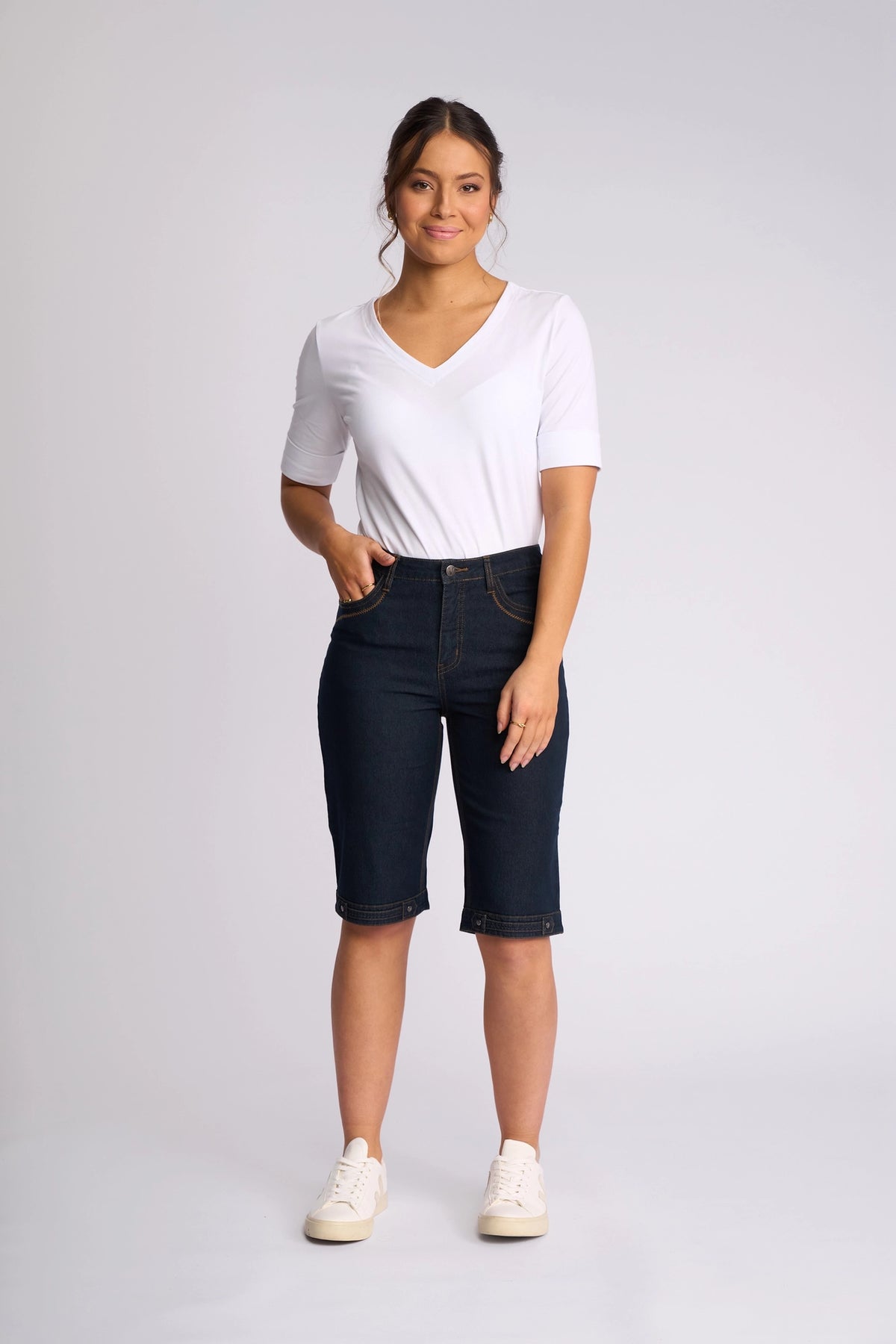 A woman stands and smiles against a plain light gray background, wearing a white V-neck T-shirt, Vassalli Custom Short in Indigo Denim—stylish stretch knee-length shorts perfect for summer—and white sneakers.