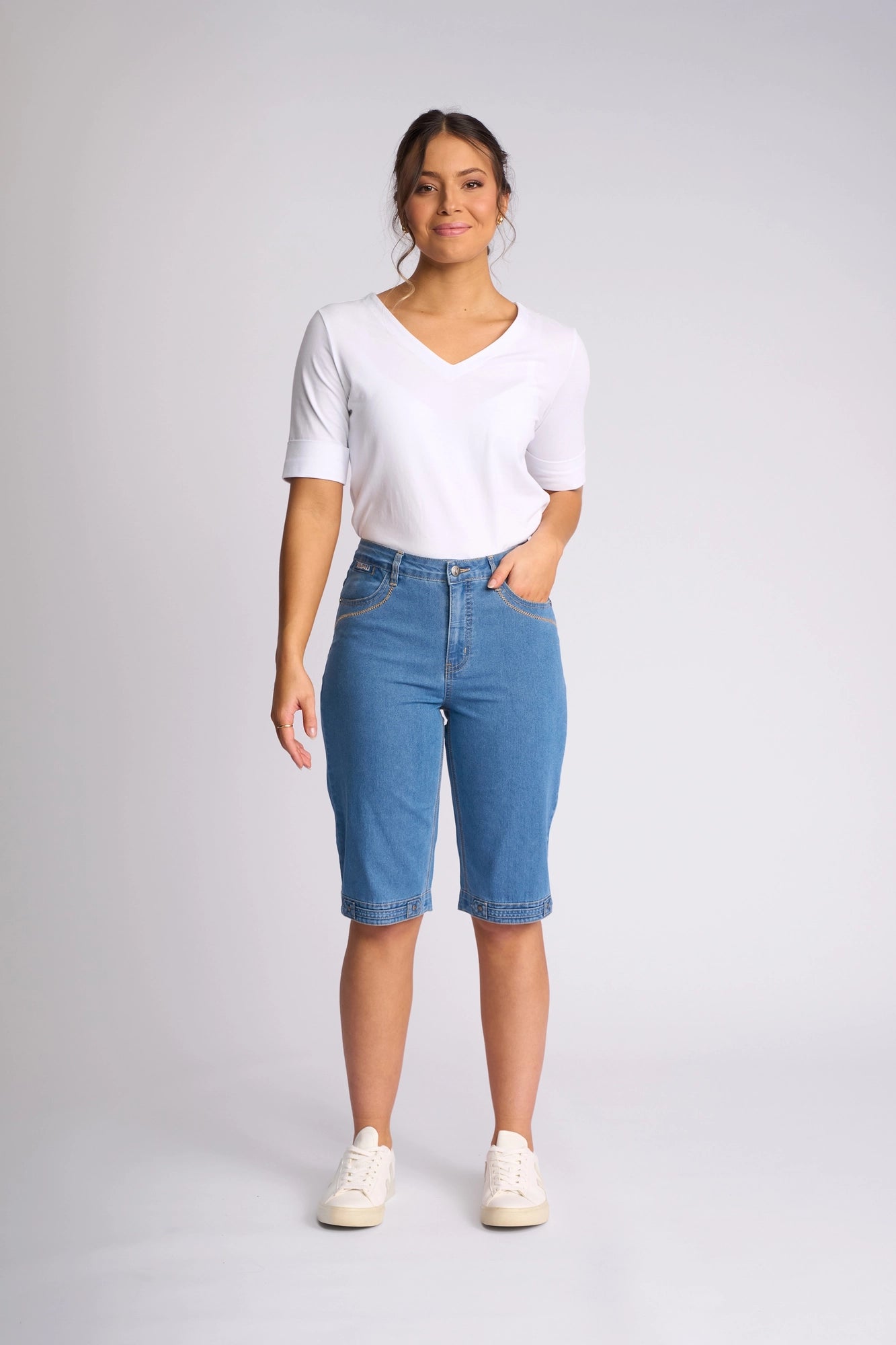 A person stands against a plain background, wearing a white short-sleeve V-neck shirt, Vassalli Custom Short - Light Denim, and white sneakers, with one hand in their pocket and a slight smile.