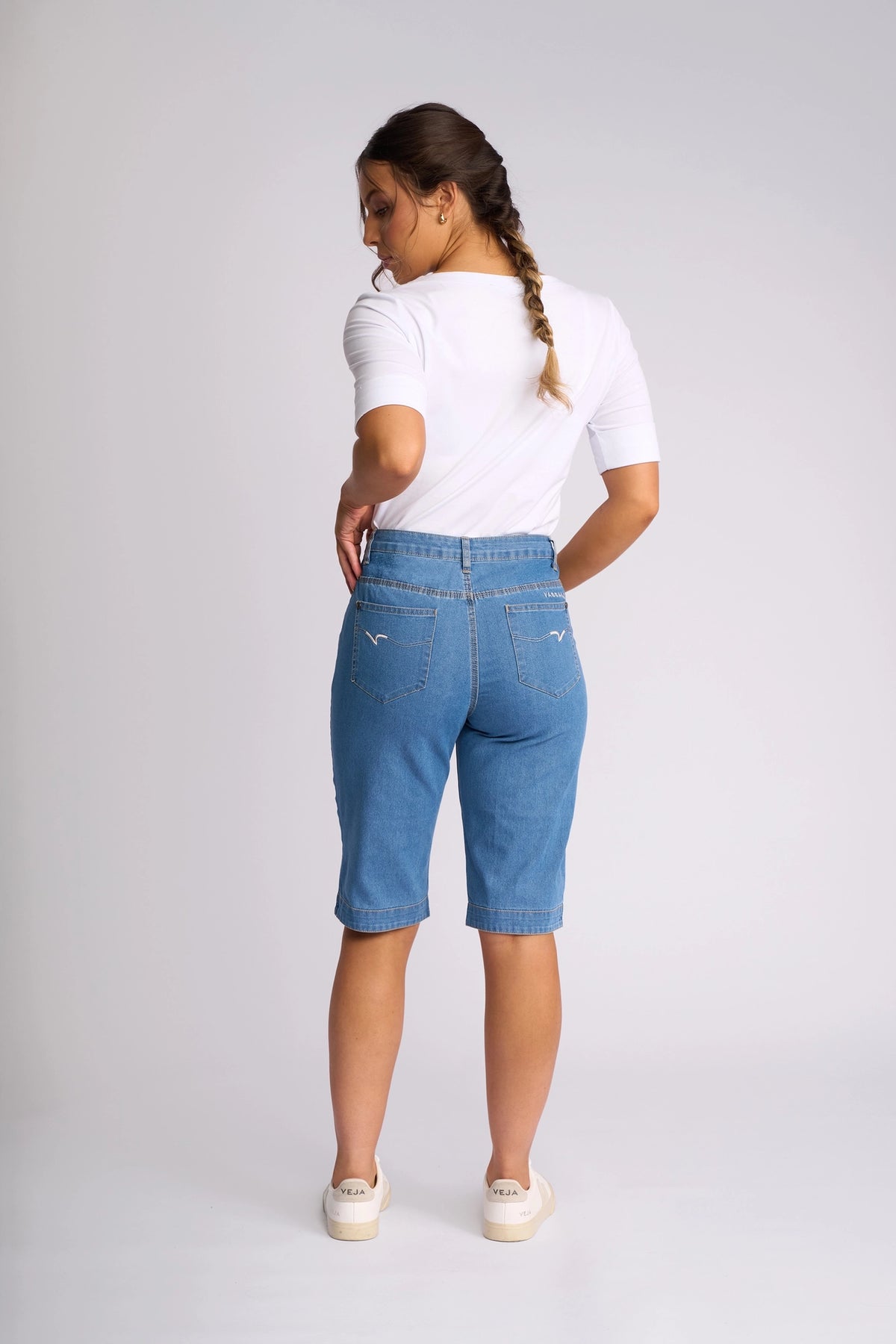 A woman with a braid, wearing a white shirt, Vassalli Custom Short - Light Denim that reach her knees, and white shoes, stands facing away in a plain studio setting.