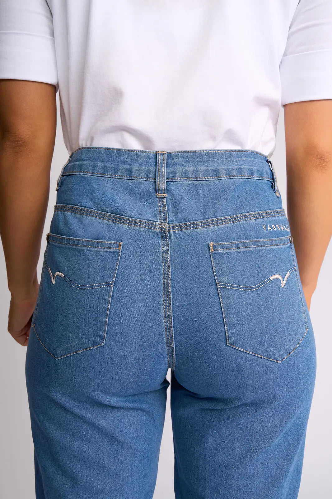A person is shown from the back wearing Vassalli’s Custom Short - Light Denim. These high-waisted, light wash blue denim shorts feature stretch fabric, two back pockets with distinctive white stitching, and a small Vassalli logo on the right pocket, paired with a white shirt.