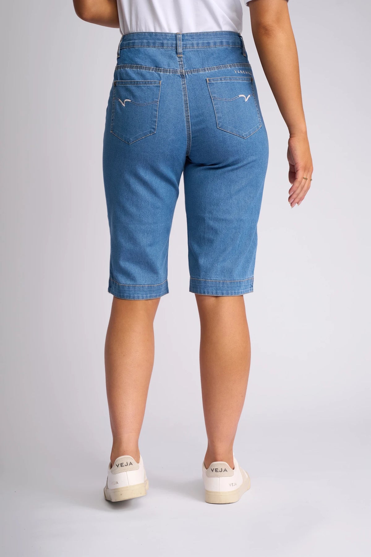 A person wearing a white shirt, Vassalli Custom Short - Light Denim, and white Veja sneakers is standing with their back to the camera against a light gray background.