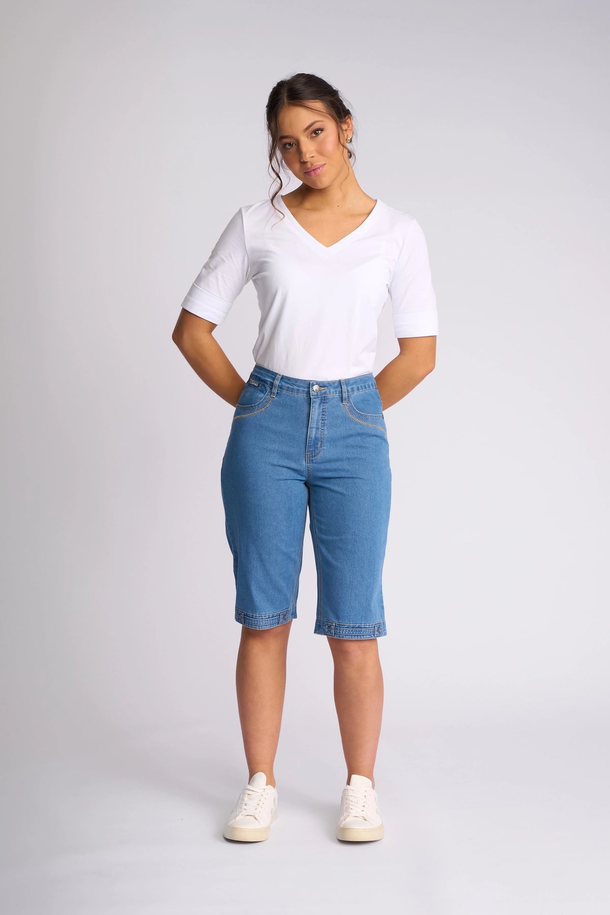 A woman stands against a plain background wearing a white V-neck shirt, white sneakers, and the Vassalli Custom Short - Light Denim, with her hands in her pockets and a neutral expression.