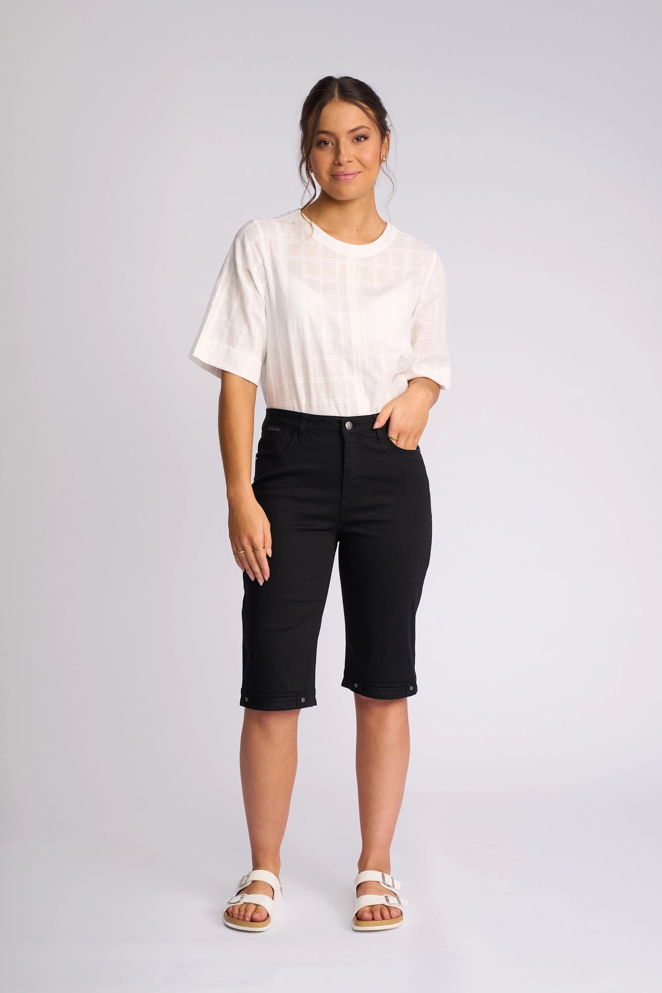 A woman stands against a plain background wearing a white short-sleeve blouse, the Vassalli Custom Lightweight Short in black, and white sandals. She has a relaxed posture and a neutral expression.