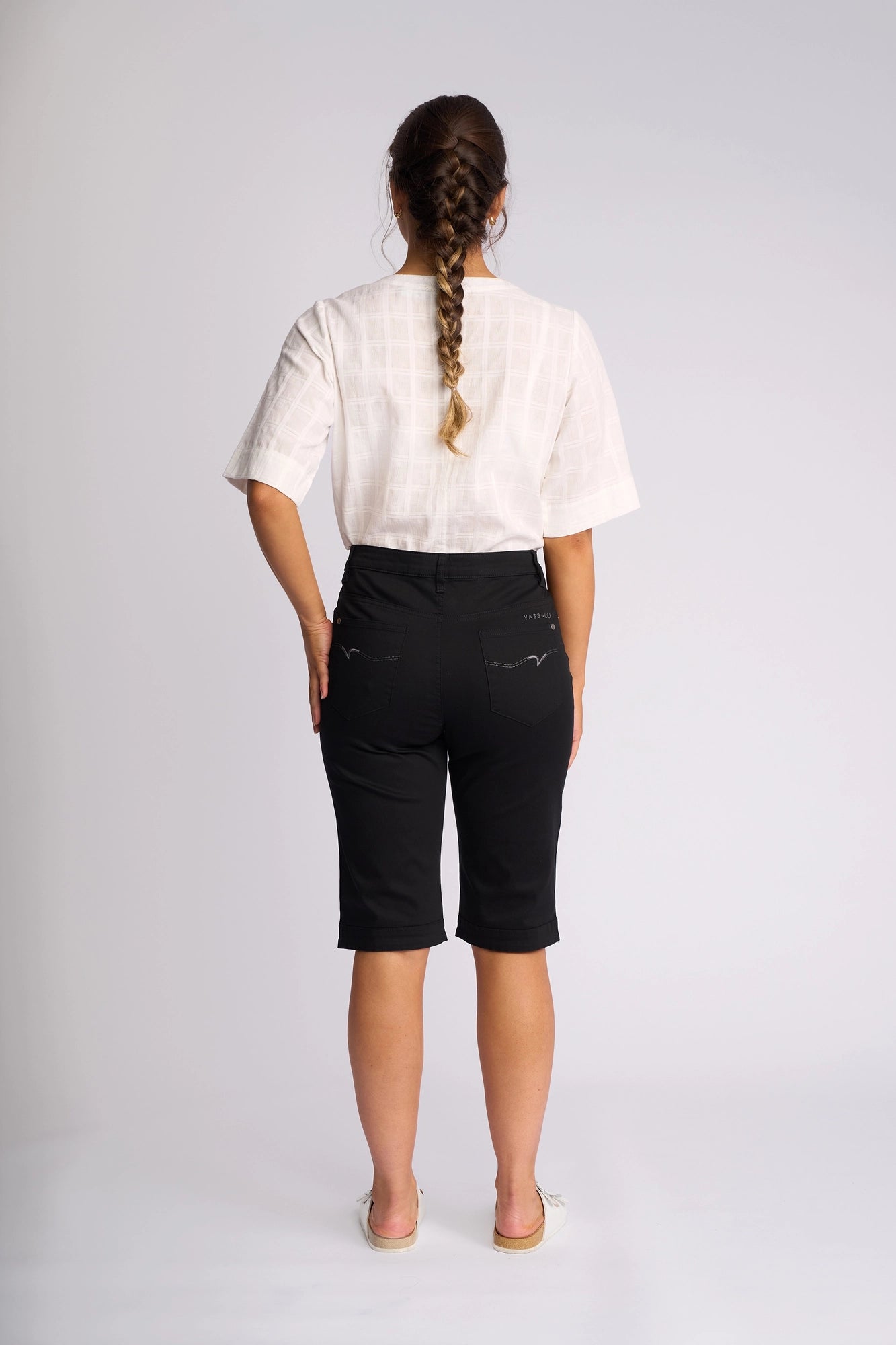 A person with a braid, dressed in a white short-sleeve shirt, the Vassalli Custom Lightweight Short in black, and white shoes stands facing away against a plain light background.