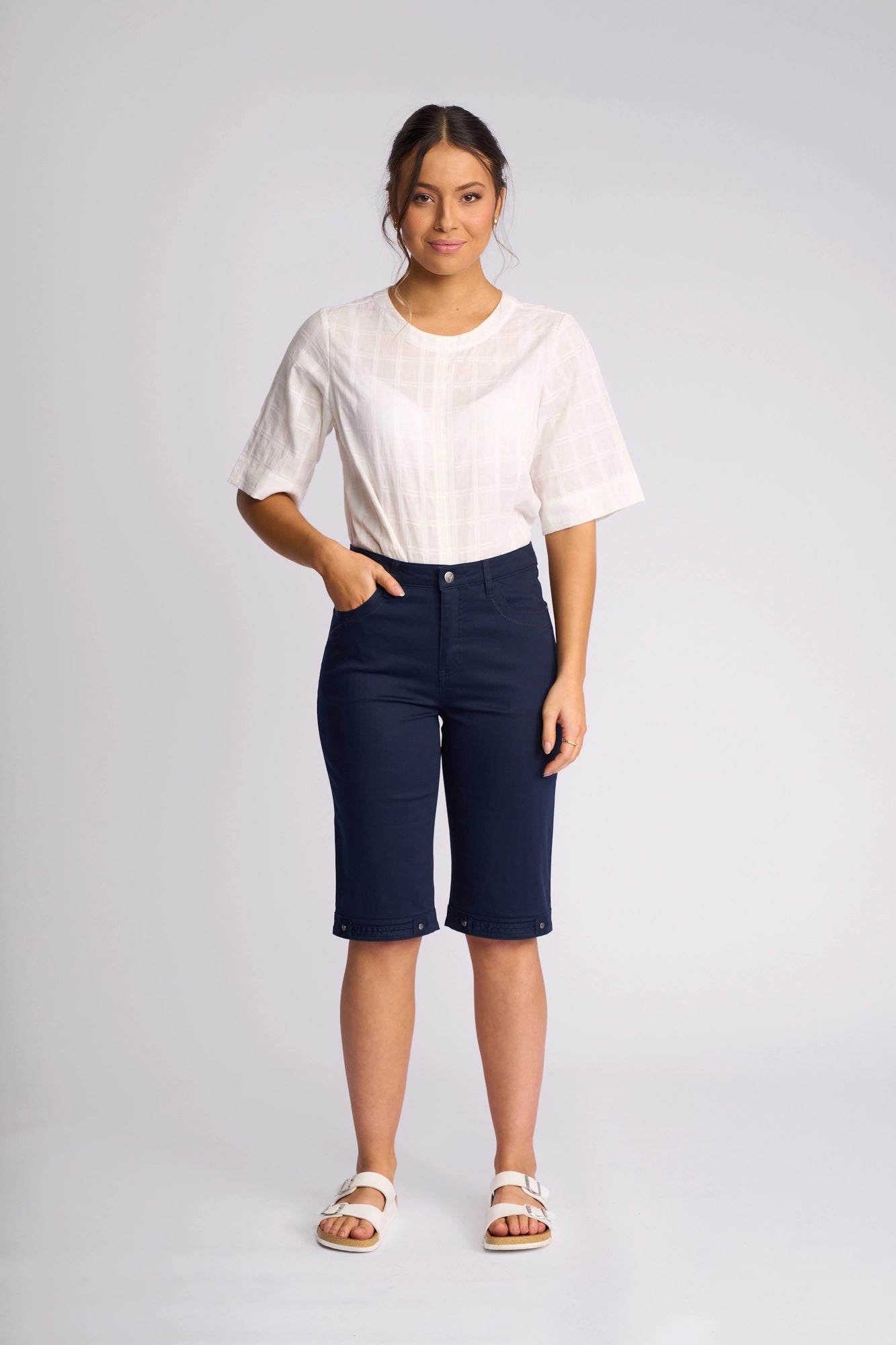 A woman stands against a plain background wearing a white short-sleeve blouse, Vassalli Custom Lightweight Short in navy crafted from comfortable cotton-elastane, and white sandals, with one hand in her pocket and a slight smile on her face.