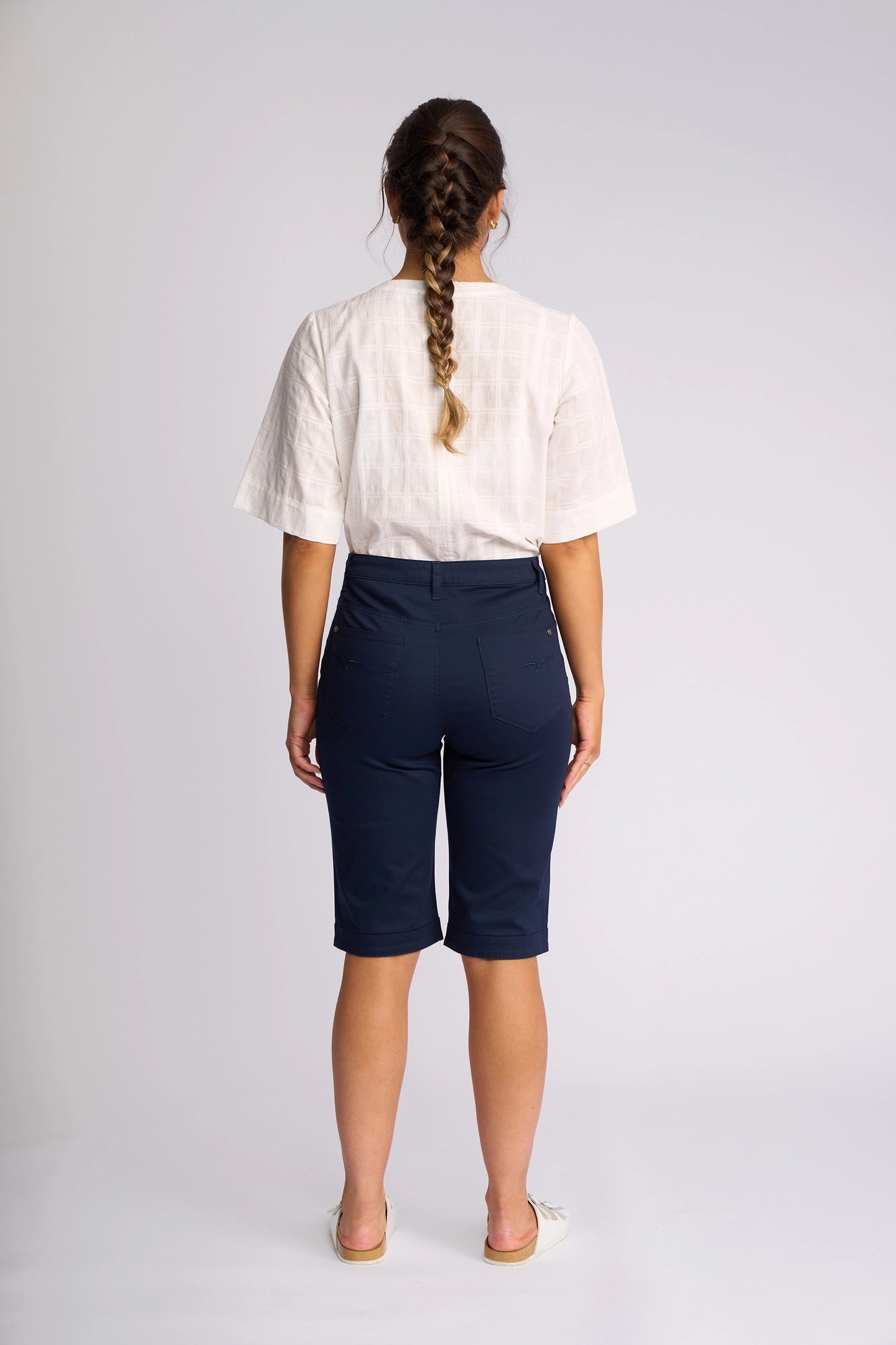 A woman with a braid stands facing away against a plain light background, wearing a white short-sleeve top, white shoes, and Vassalli’s Custom Lightweight Short in navy.