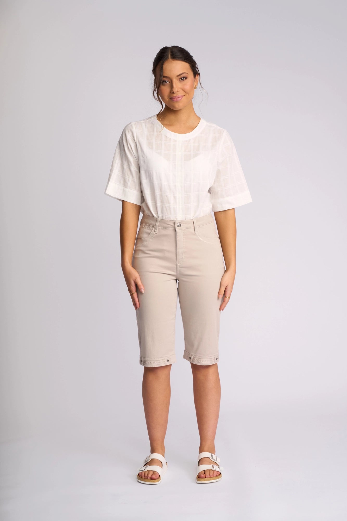 A woman stands facing the camera, wearing a white short-sleeve blouse paired with Vassalli’s Custom Lightweight Short in Pumice, styled with white sandals against a plain light background—a perfect look for effortless summer dressing.
