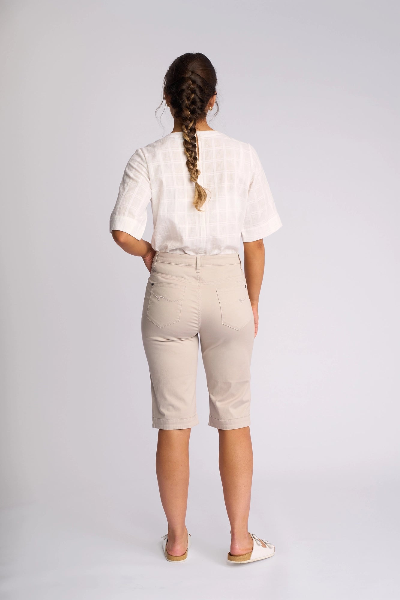 A woman with braided hair stands facing away, wearing a white short-sleeve shirt, Vassalli Custom Lightweight Short in Pumice—crafted from soft cotton-elastane for effortless summer dressing—and white slip-on shoes against a plain light background.
