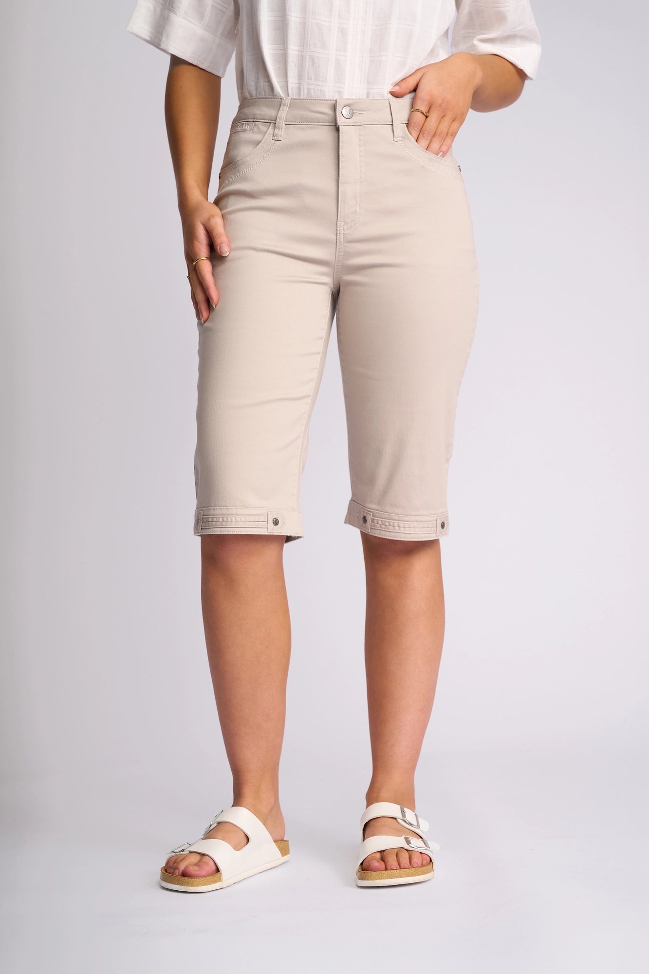 Wearing a white shirt, Vassalli’s Custom Lightweight Short in Pumice—a beige, knee-length style crafted from breathable cotton-elastane fabric—and white sandals, this person stands against a plain background for an effortless summer look.