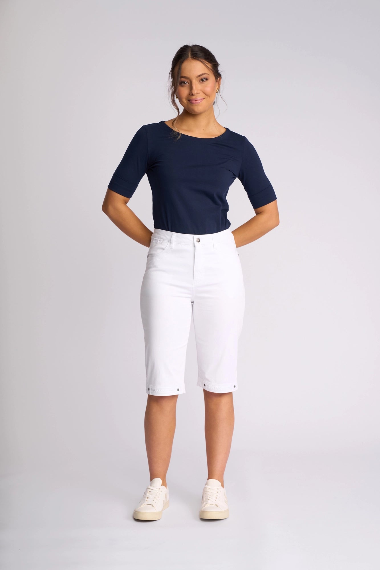 A woman stands against a plain background, wearing the Vassalli Custom Lightweight Short in white—crafted from breathable cotton-elastane—and a navy blue short-sleeve top, paired with white sneakers. With her hands behind her back and a slight smile, she exemplifies effortless summer style.