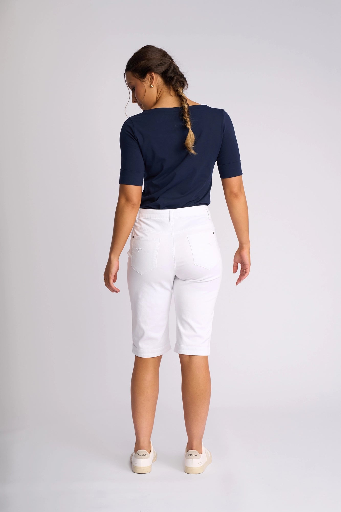 A woman with a braid, wearing a navy top, Vassalli’s Custom Lightweight Short in white—crafted from cotton-elastane for perfect summer dressing—and white sneakers, stands facing away against a plain light background.
