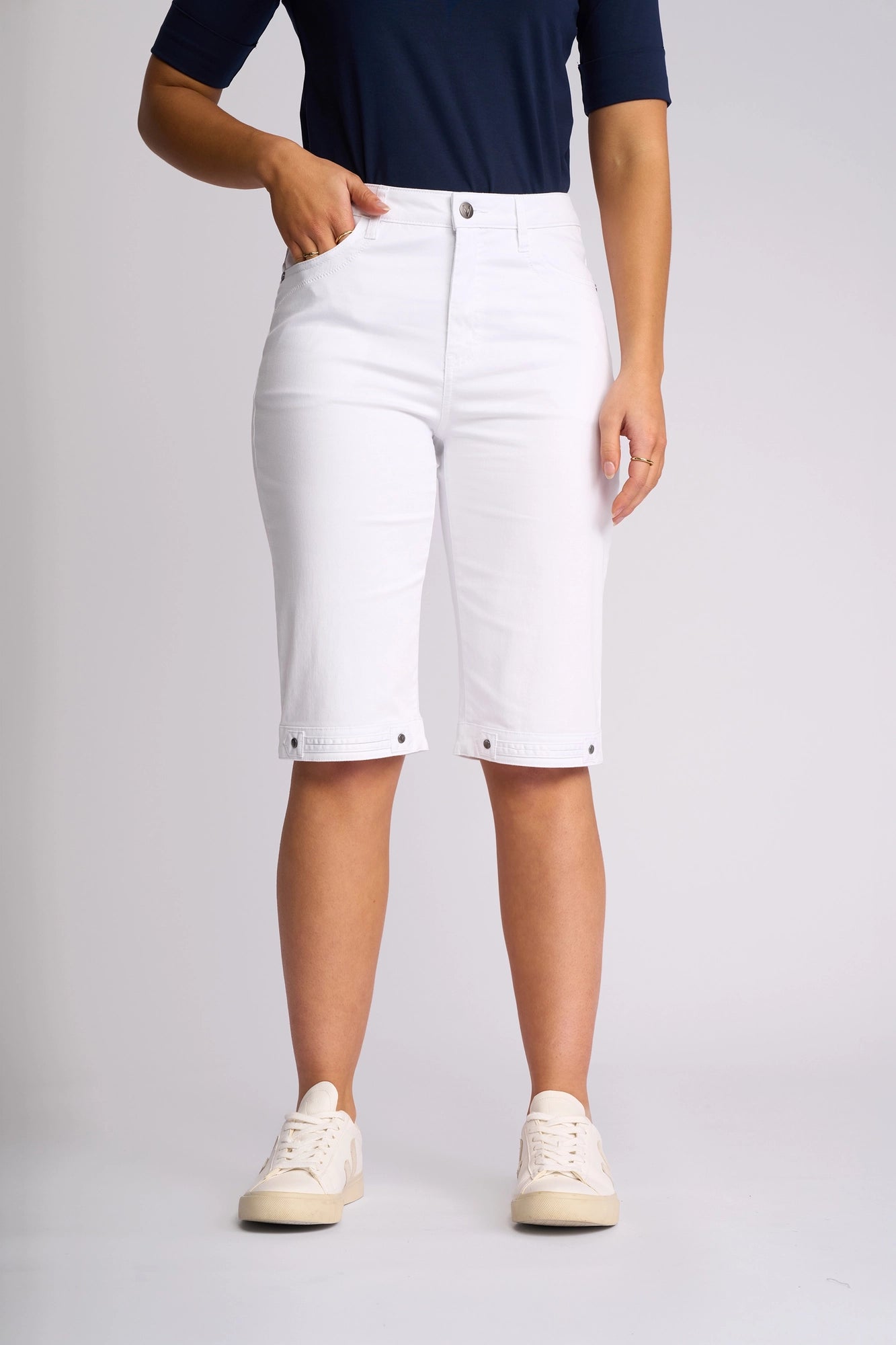 A person wearing a dark blue shirt, Vassalli Custom Lightweight Short in White—crafted from lightweight cotton with a knee-length, straight-leg fit and front pockets—and white sneakers stands against a plain light background.