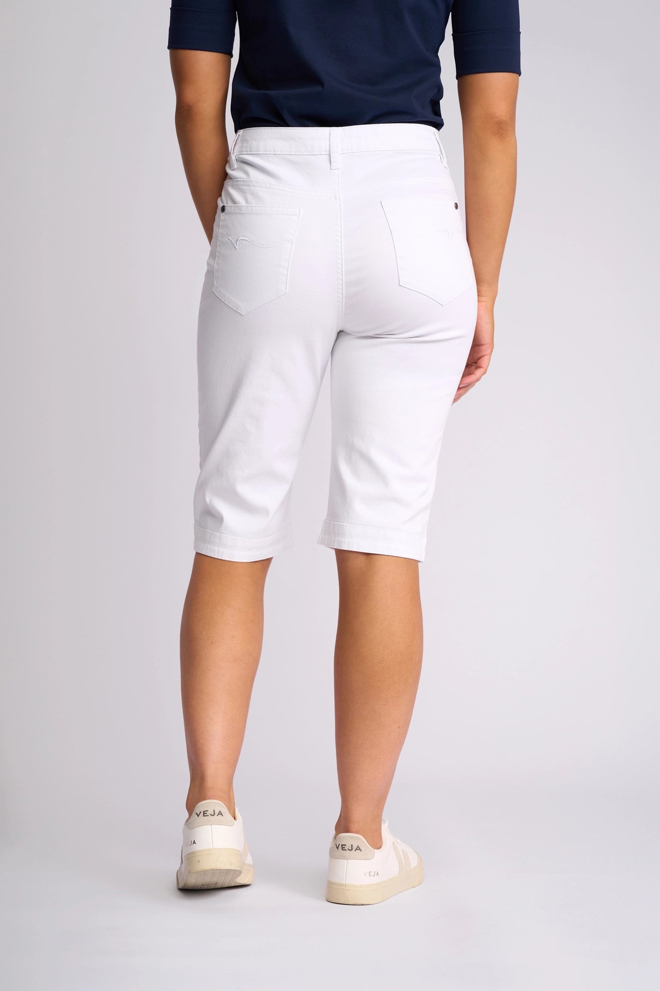 A person is standing facing away against a plain light background, wearing Vassalli’s Custom Lightweight Short in white—crafted from lightweight cotton with a straight-leg fit—paired with a navy-blue shirt and white Veja sneakers.