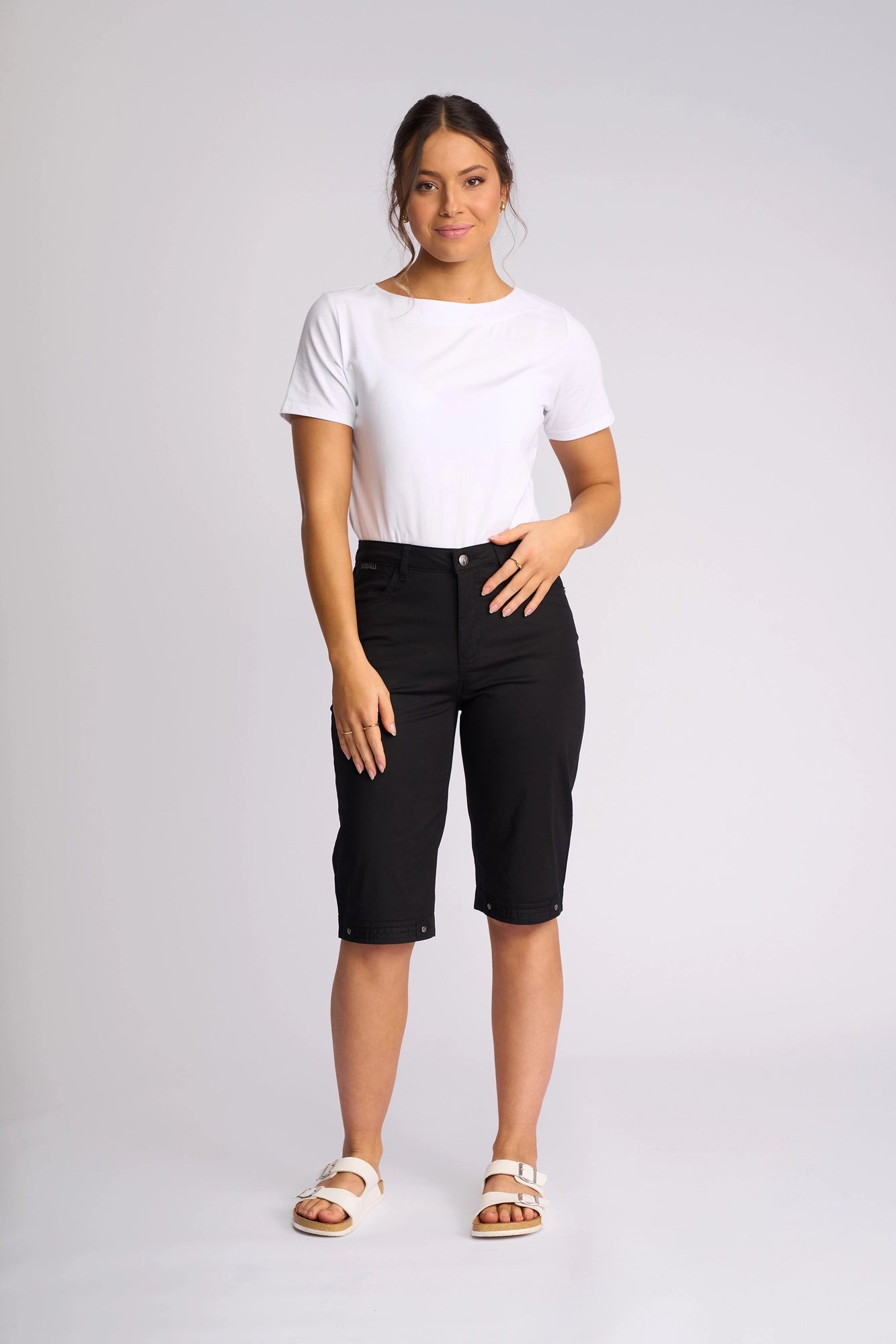 A woman stands facing forward against a plain light background wearing the Vassalli Custom Poplin Short in black—a lightweight summer staple—paired with a white t-shirt and white sandals.