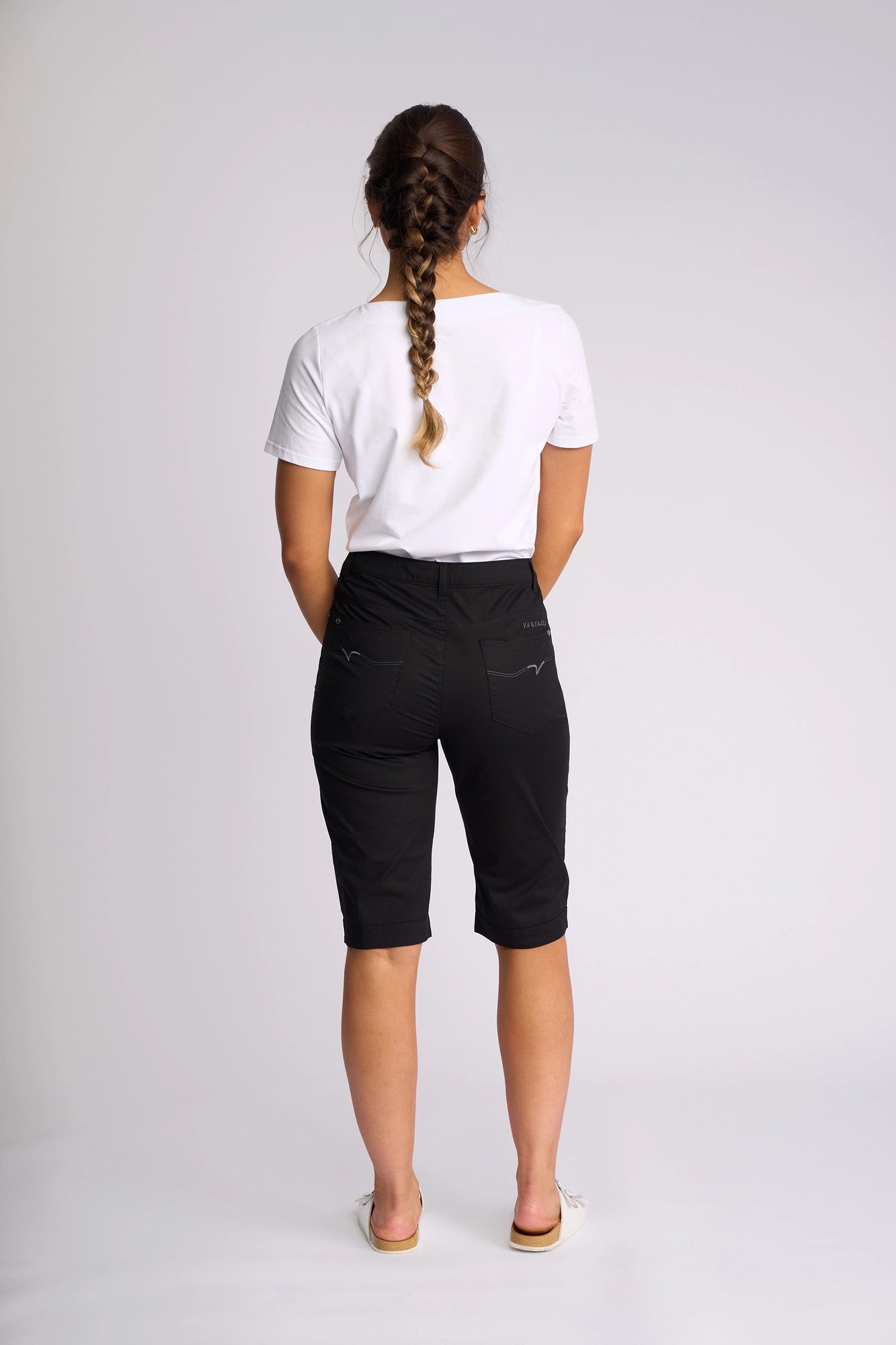 A person with a long braid, dressed in a white t-shirt and Vassalli’s Custom Poplin Short in black—a lightweight summer staple—paired with flip-flops, stands facing away against a plain light background.
