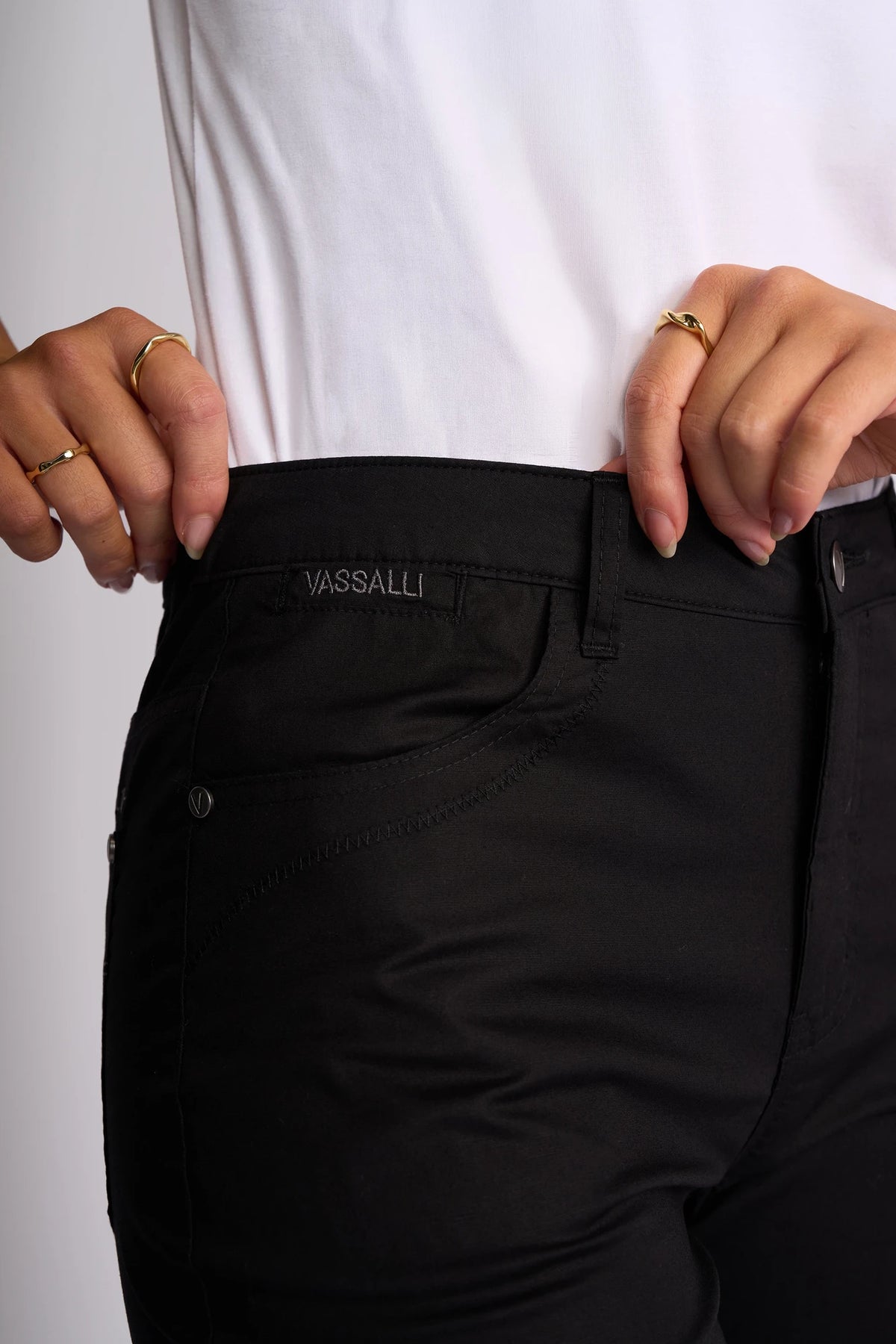 A person wearing the Vassalli Custom Poplin Short in black is shown from the waist down, holding the waistband. Gold rings adorn their fingers, drawing attention to this lightweight summer staple.