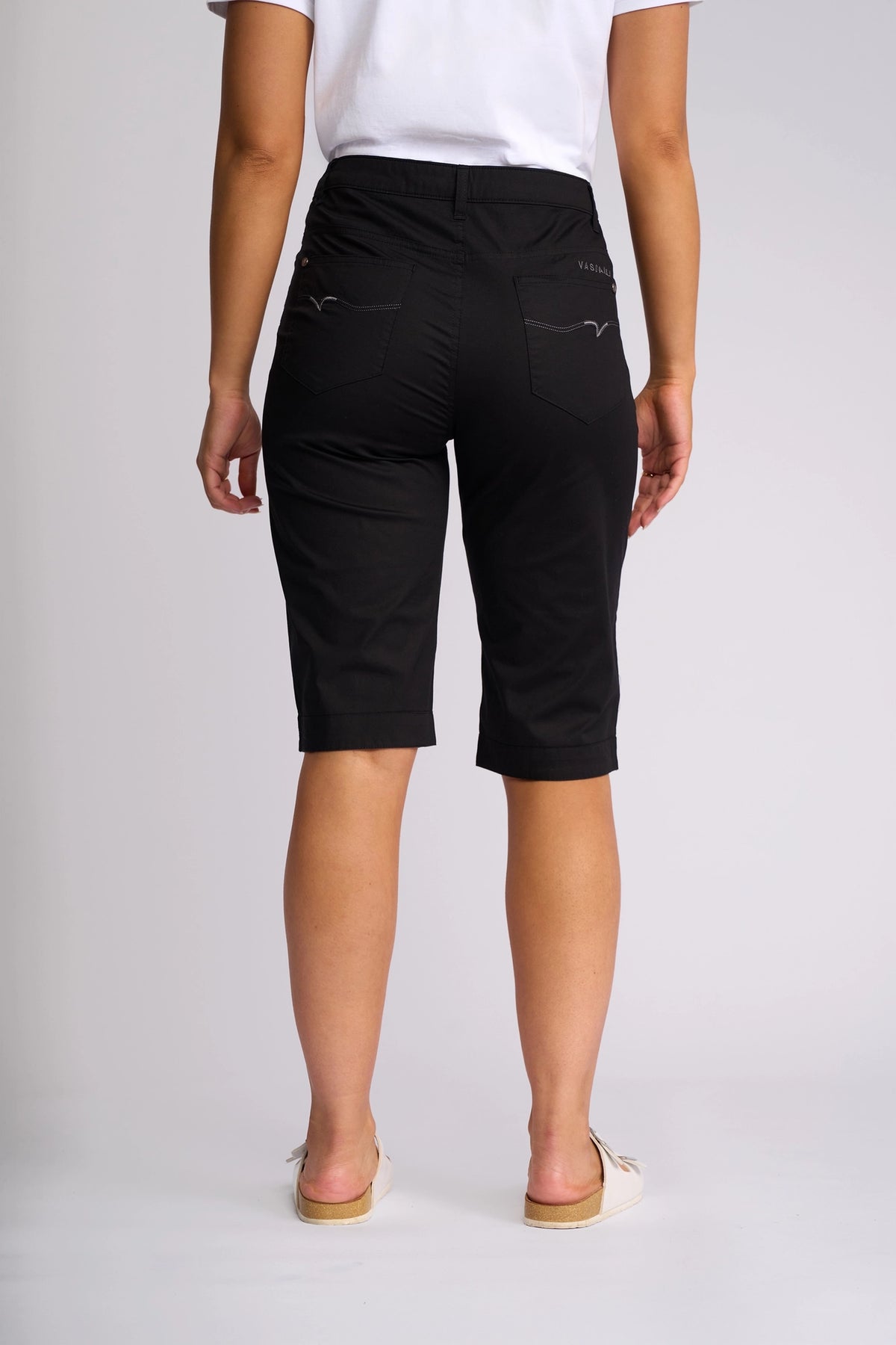 Seen from behind, a person wears a white shirt and the Vassalli Custom Poplin Short in black—an effortlessly lightweight summer staple—paired with white slip-on shoes against a plain background.