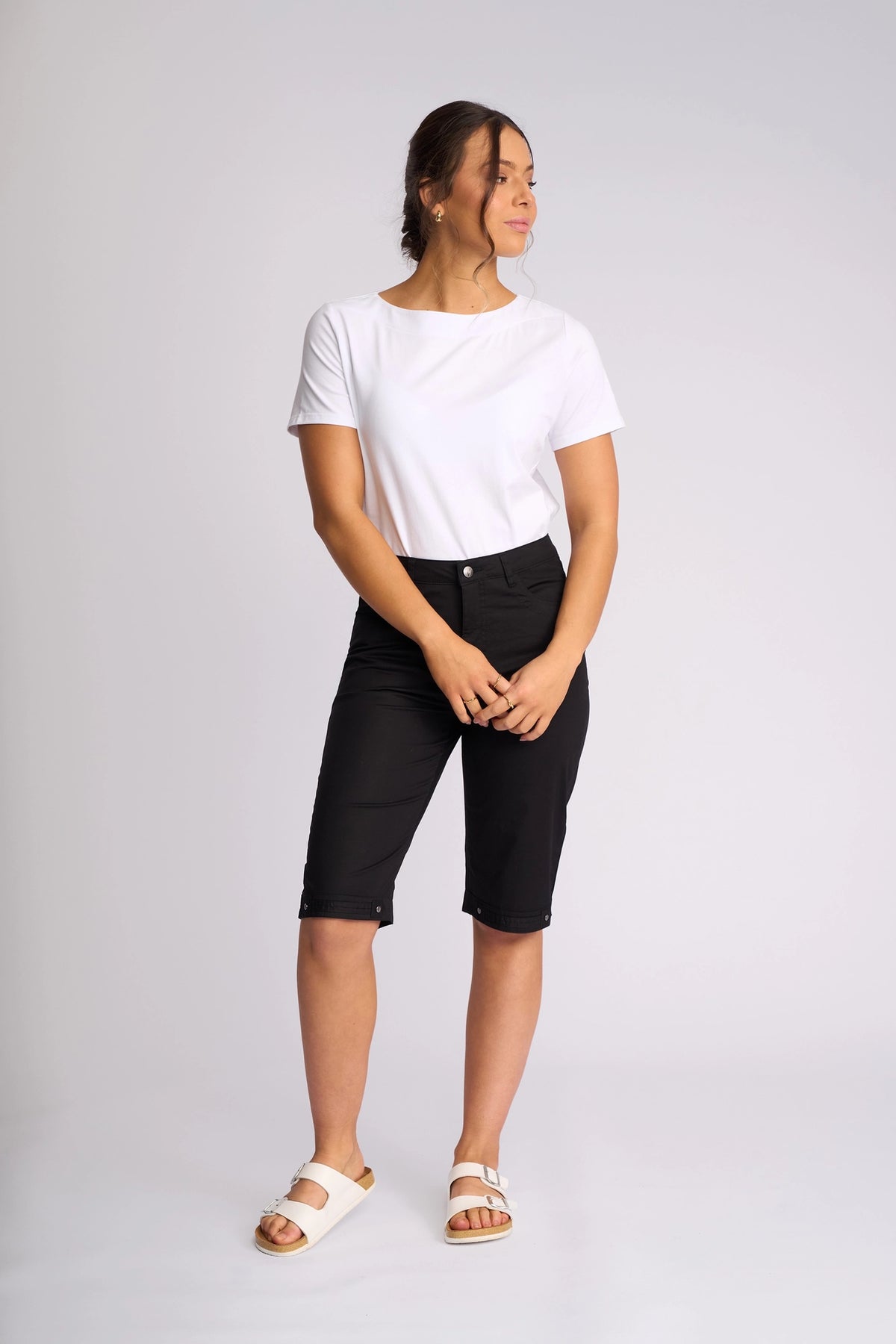 A woman stands against a plain background wearing a white t-shirt, the Vassalli Custom Poplin Short in black—an effortlessly lightweight summer staple—and white sandals. She has her hands clasped in front of her and is looking slightly to the side.