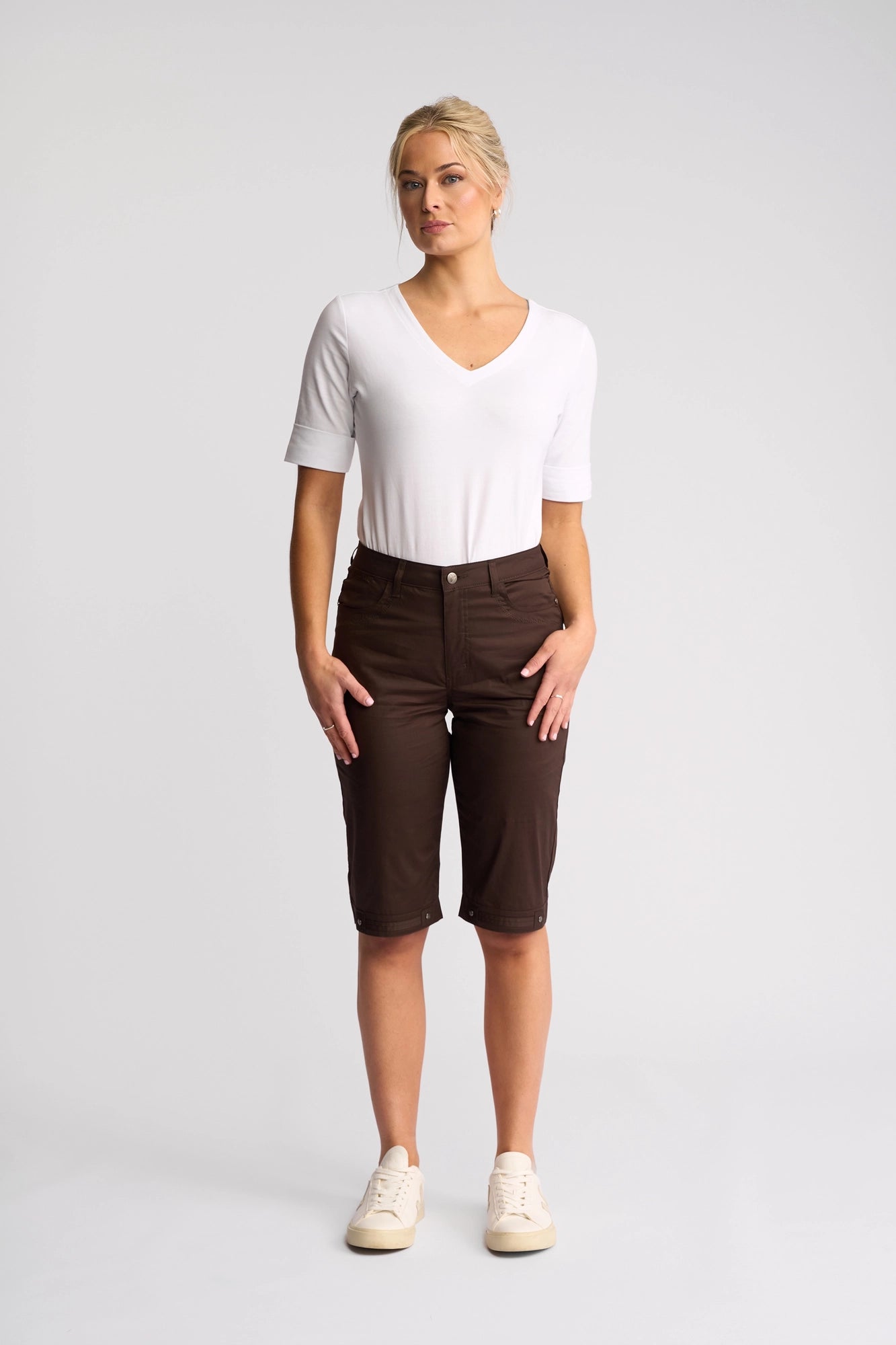 A woman stands facing forward, wearing a white short-sleeve V-neck top, Vassalli’s Custom Poplin Short in Chocolate—an effortless summer staple—and beige sneakers, against a plain light gray background.