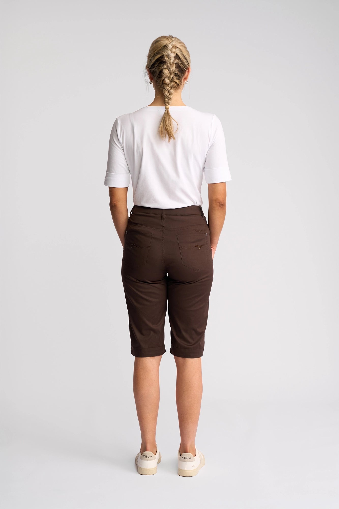 A person with blonde hair in two braids, wearing a white t-shirt and the Vassalli Custom Poplin Short in Chocolate—a true summer staple—paired with white shoes, stands facing away against a plain light background.