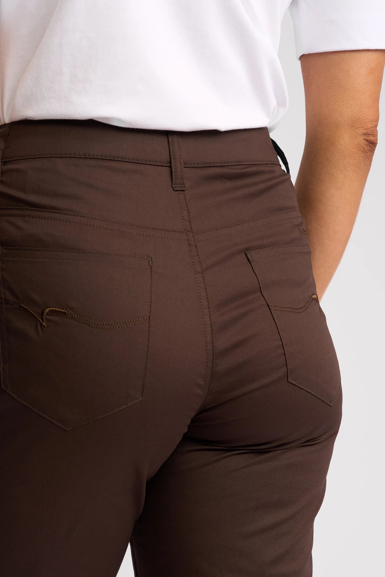 A person is shown from the back waist down wearing Vassalli Custom Poplin Short in Chocolate and a white shirt. The summer staple shorts feature two back pockets with visible stitching, and the person’s right arm is bent slightly at the elbow.