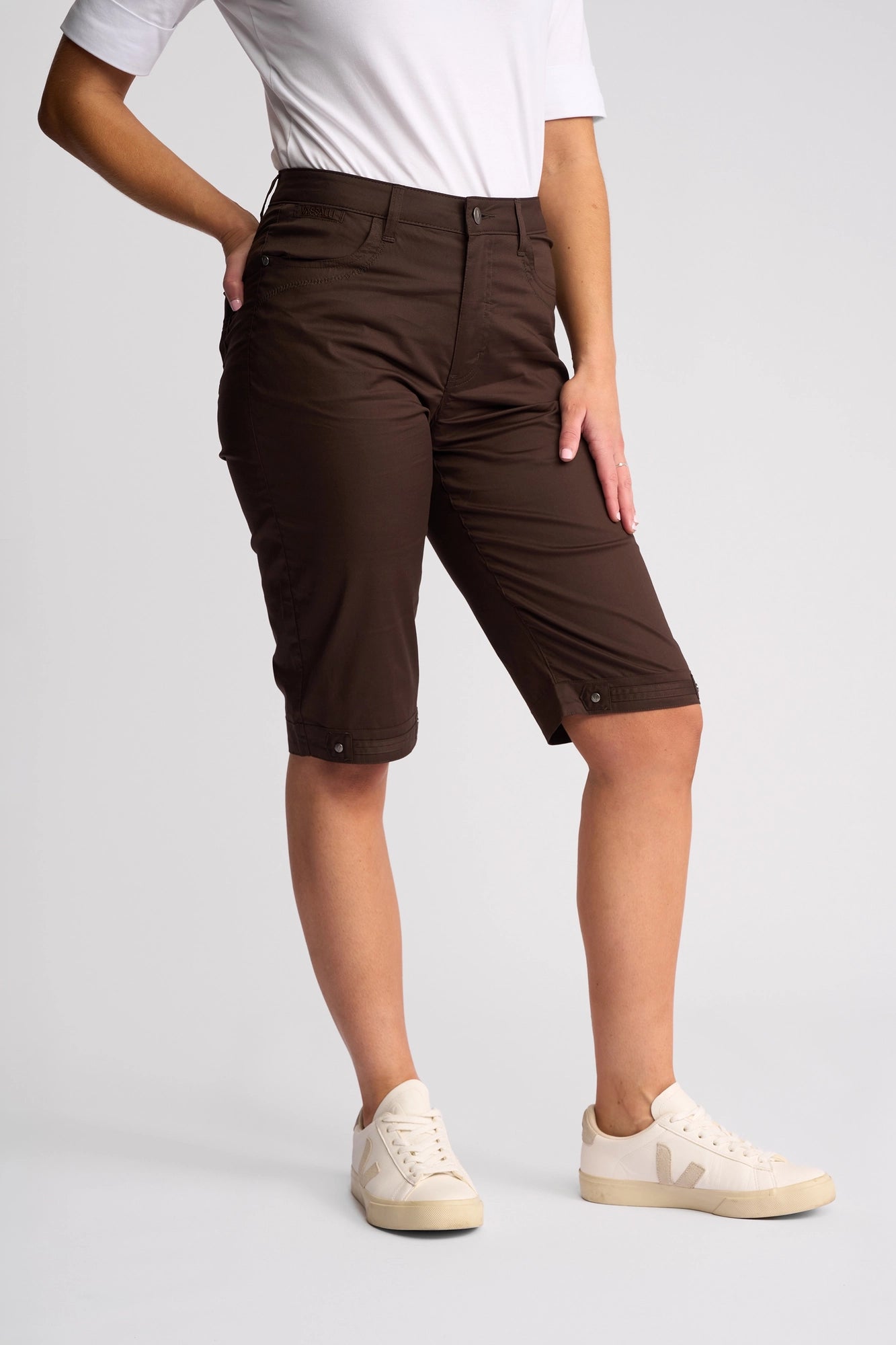 A person wearing a white t-shirt, Vassalli’s Custom Poplin Short in Chocolate—styled in a straight-leg cut—and white sneakers stands with one hand on their hip against a plain background, embodying the perfect summer staple look.