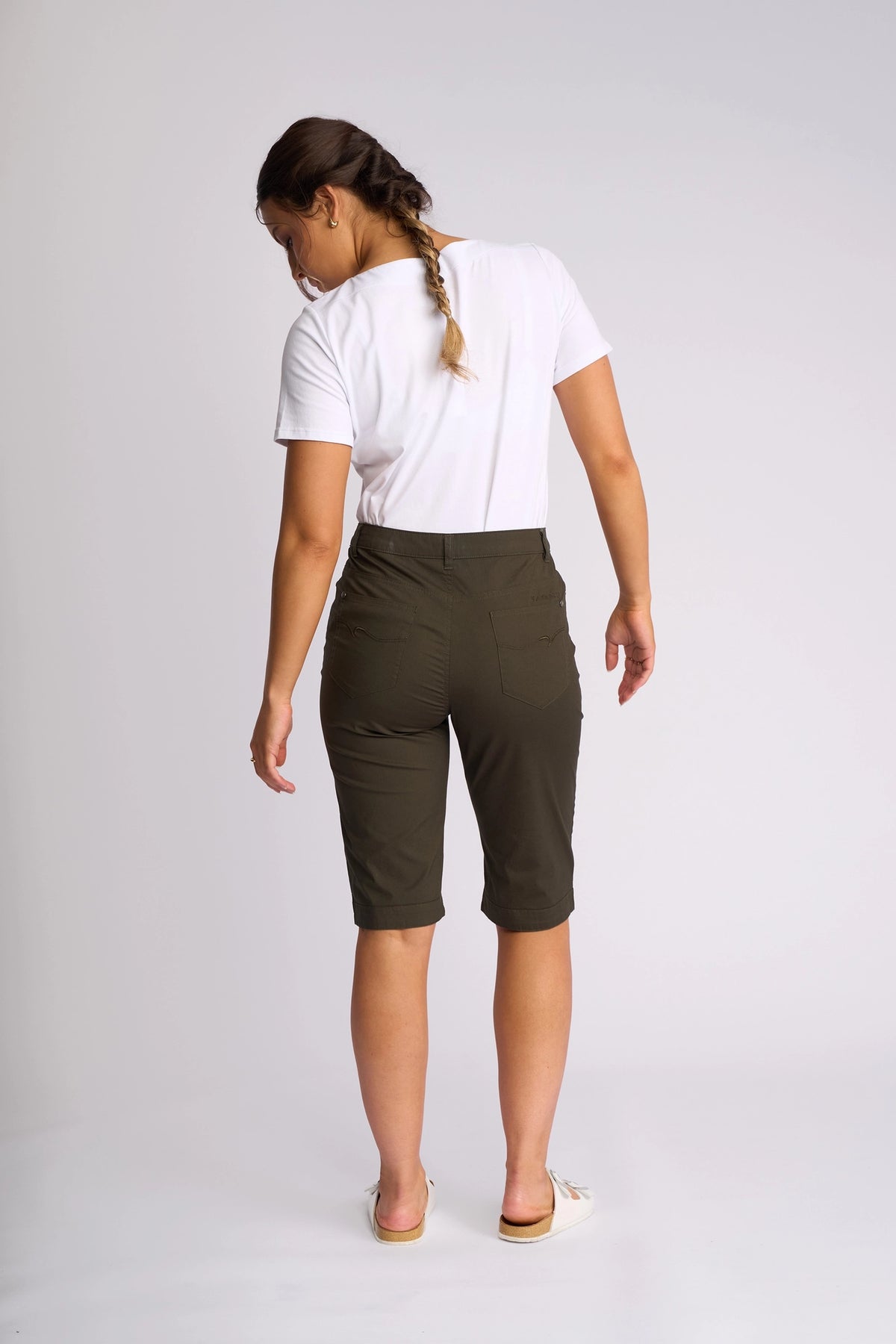 A woman with a braid, barefoot and facing away from the camera against a plain light background, wears a white t-shirt paired with Vassalli’s Custom Poplin Short in khaki.