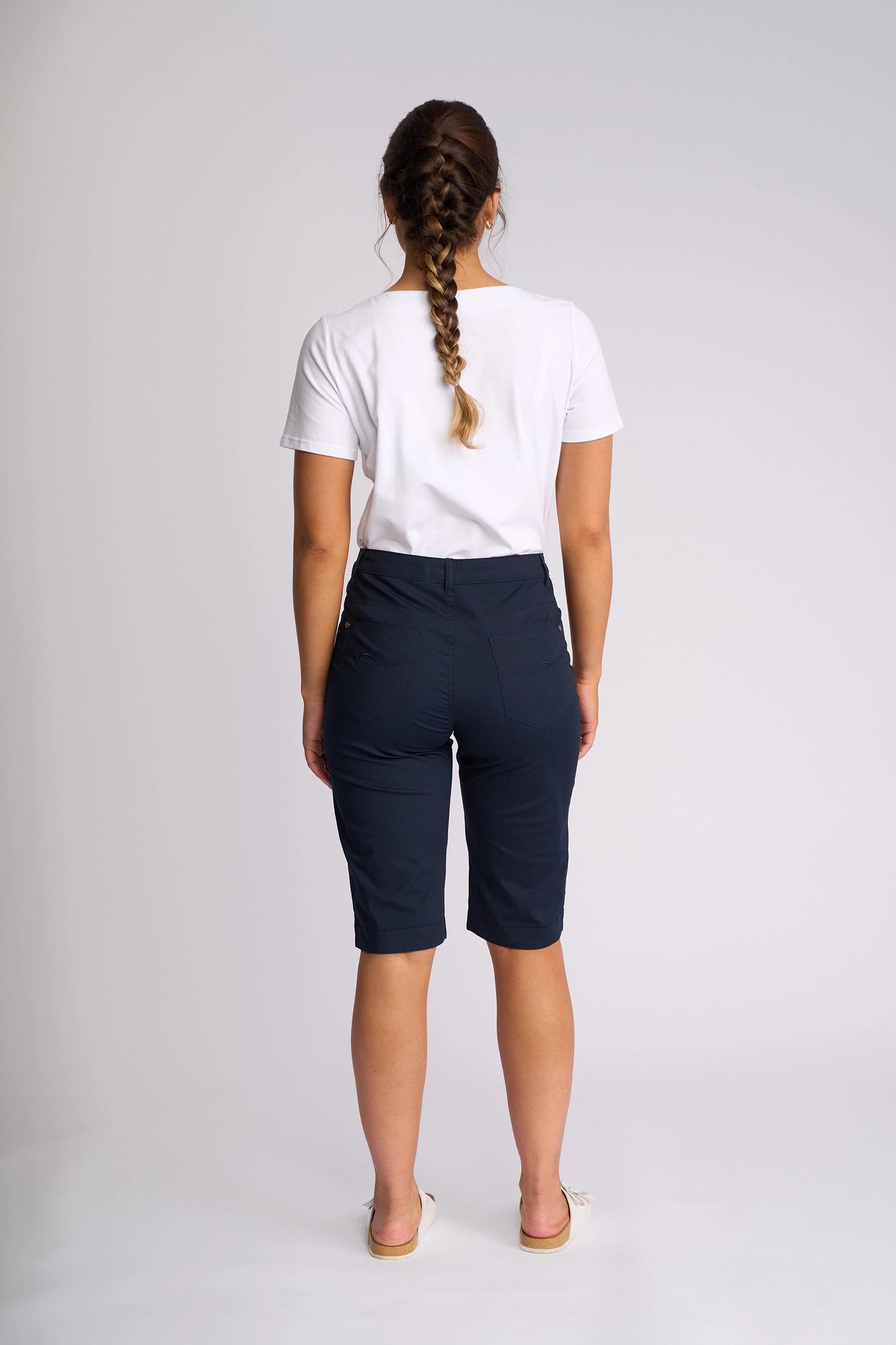 A woman with a long braid stands facing away, wearing a white t-shirt, Vassalli Custom Poplin Short in navy—a breathable summer staple—and sandals, against a plain light background.