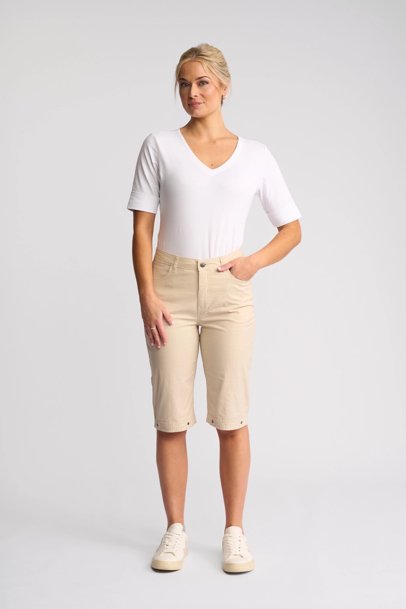 A person stands against a plain background wearing a white short-sleeve V-neck shirt, Vassalli Custom Poplin Short in Sand—an essential summer staple—and beige sneakers, with one hand in their pocket.