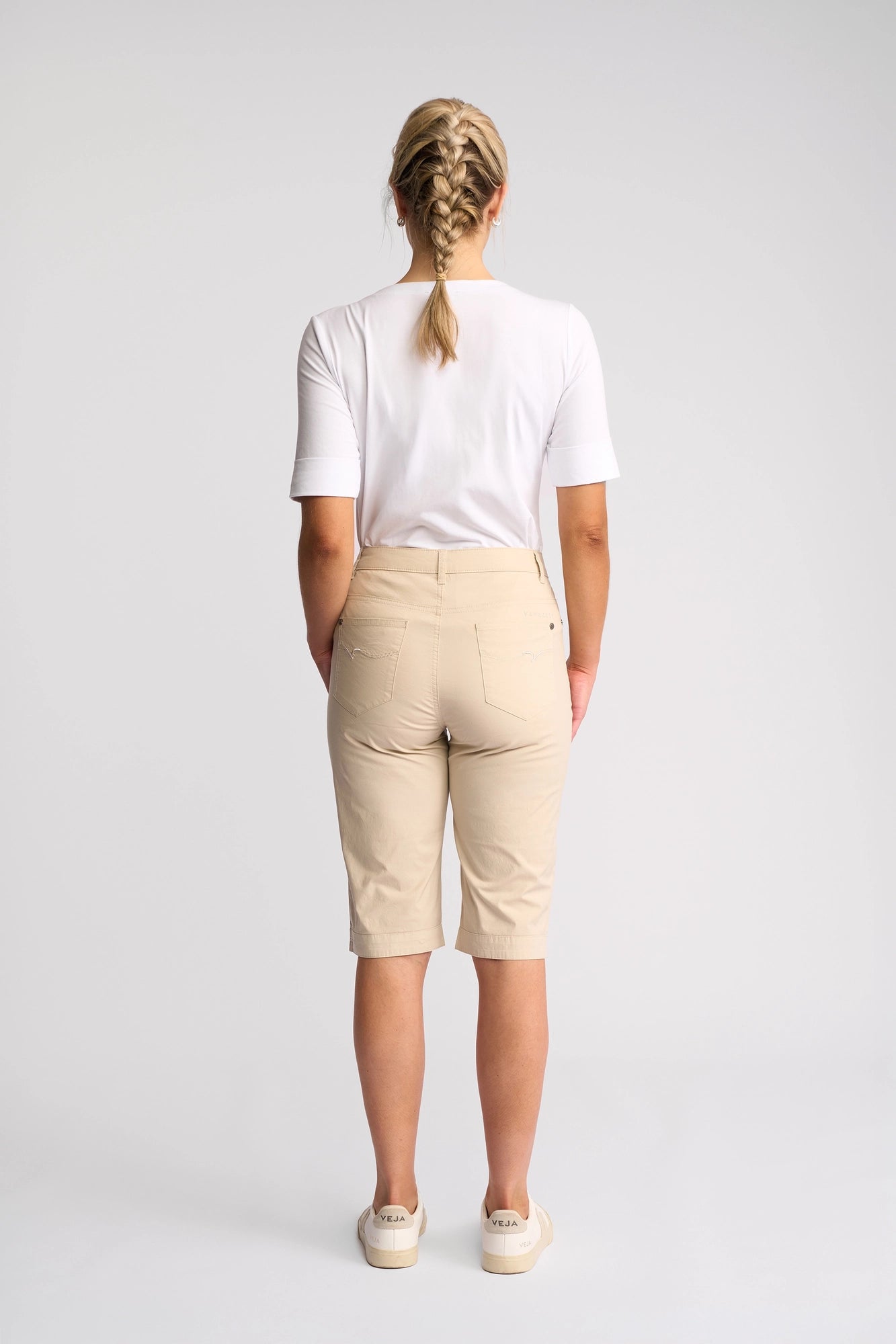 A person with braided blonde hair, dressed in a white t-shirt, Vassalli Custom Poplin Short in Sand—a true summer staple—and white shoes, stands facing away from the camera against a plain white background.