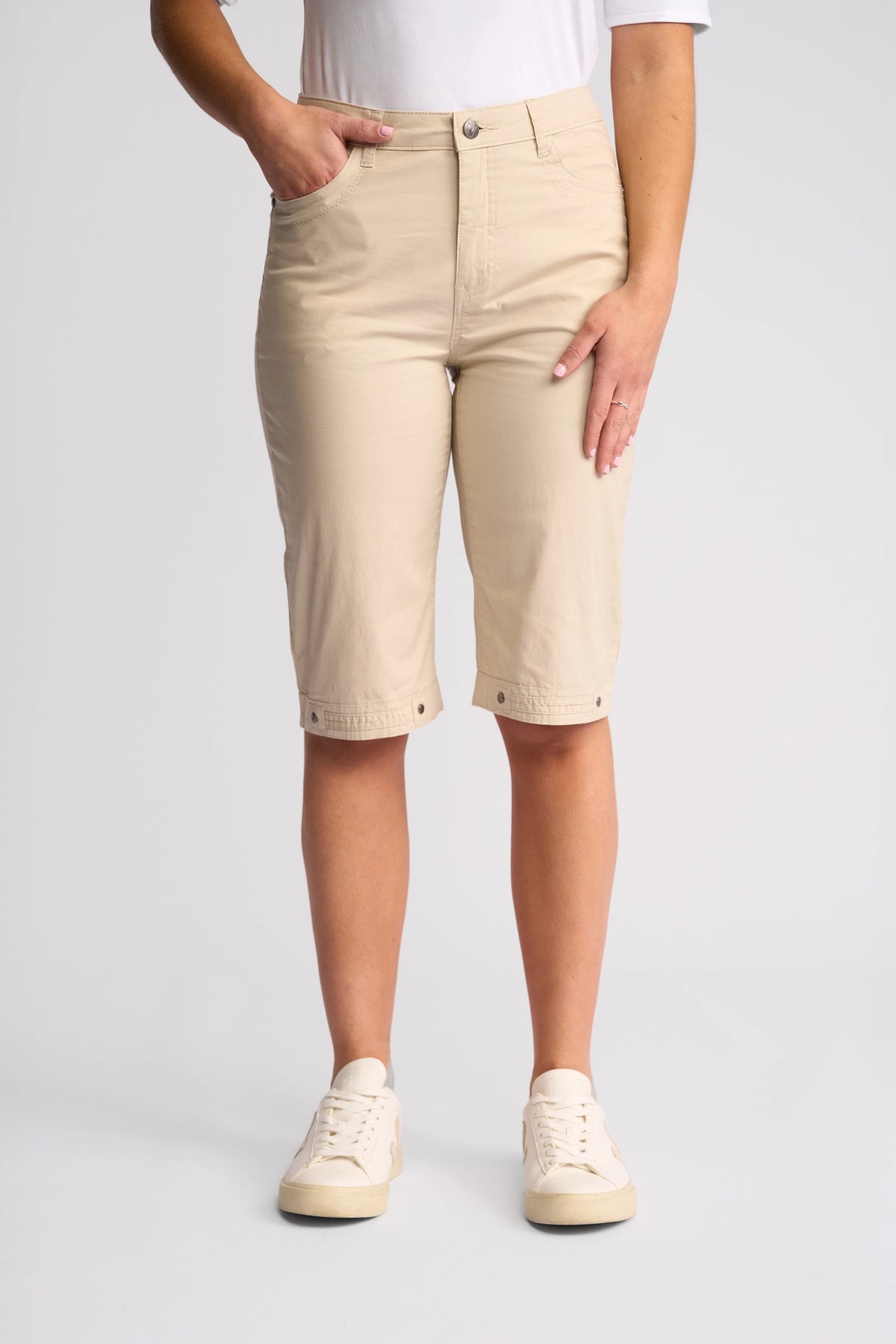 A person wearing a white shirt, Vassalli Custom Poplin Short - Sand, and white sneakers stands against a plain light background with one hand in their pocket and the other resting on their thigh—a true summer staple.