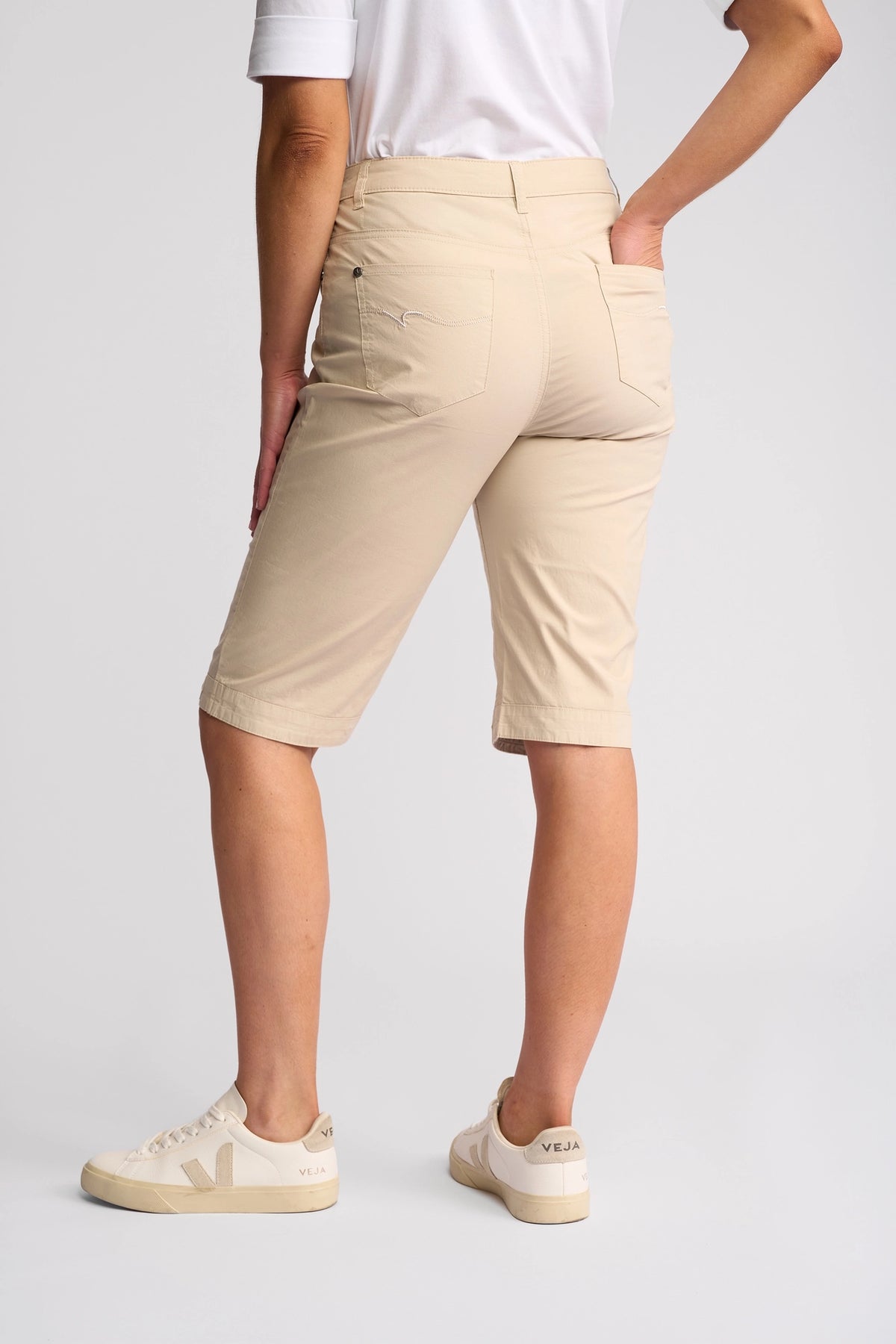 A person wearing a white shirt, Vassalli Custom Poplin Short in Sand—a true summer staple—and beige Veja sneakers stands facing away, with one hand on their hip against a plain light background.