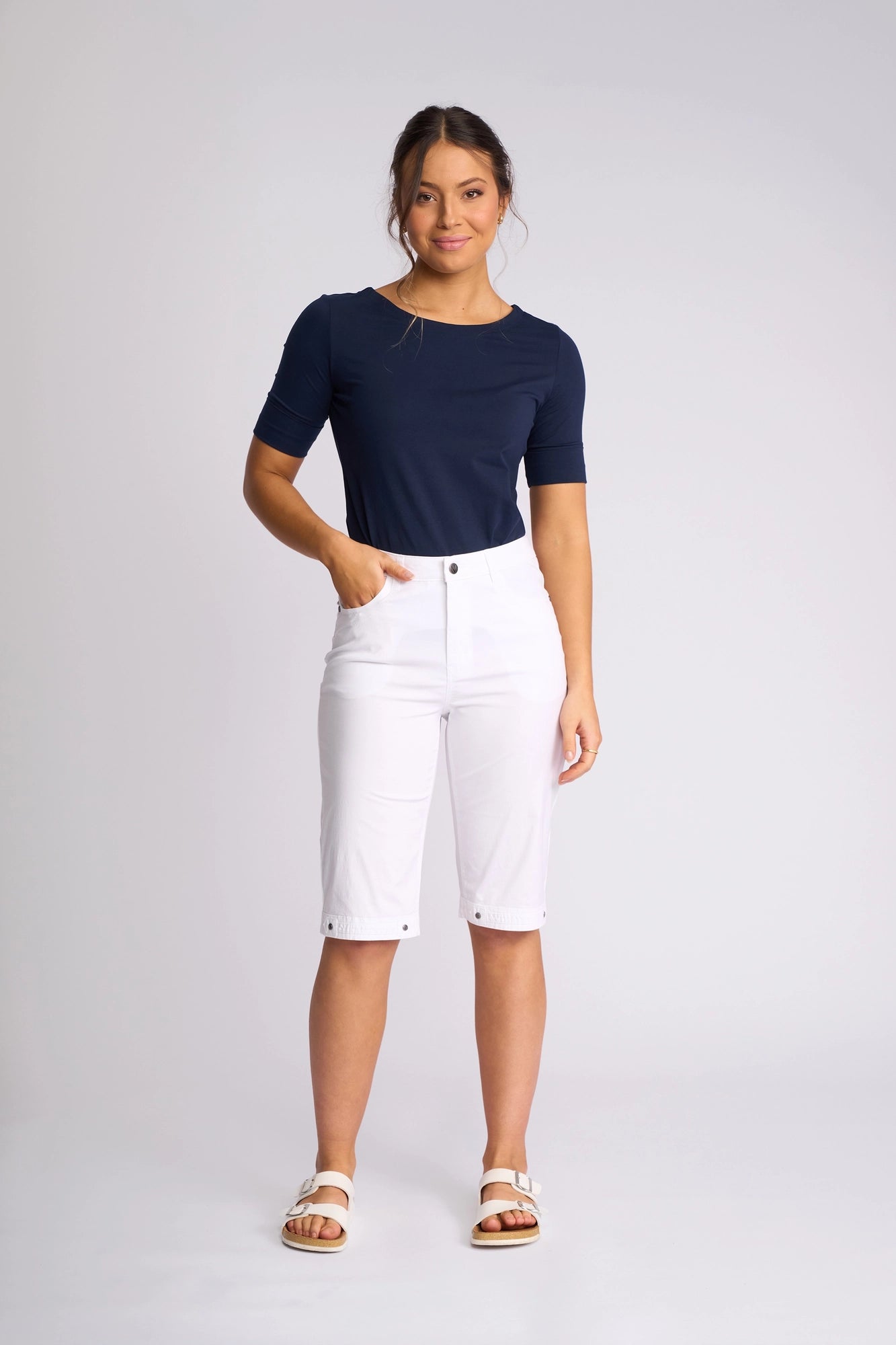 A woman stands against a plain background wearing the Vassalli Custom Poplin Short in white—an effortless summer staple—paired with a navy blue short-sleeve top and white sandals, one hand in her pocket and a slight smile on her face.