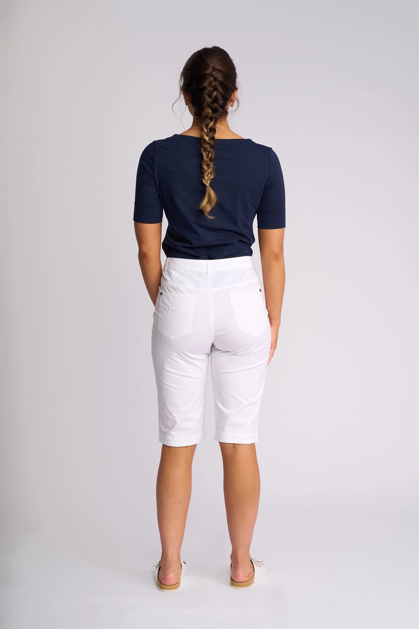 A woman stands with her back to the camera, wearing a navy short-sleeve shirt paired with Vassalli’s Custom Poplin Short in White—a classic summer staple. She completes the look with flat shoes and has her hair styled in a braid. The background is plain and light-colored.