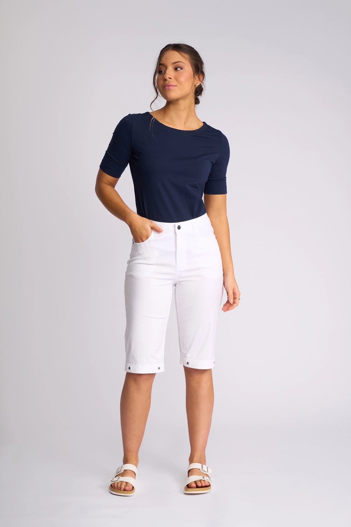 A woman stands against a plain light gray background, looking to her left with one hand in her pocket. She wears a navy blue short-sleeve top, Vassalli Custom Poplin Short in white—an essential summer staple—and white sandals.